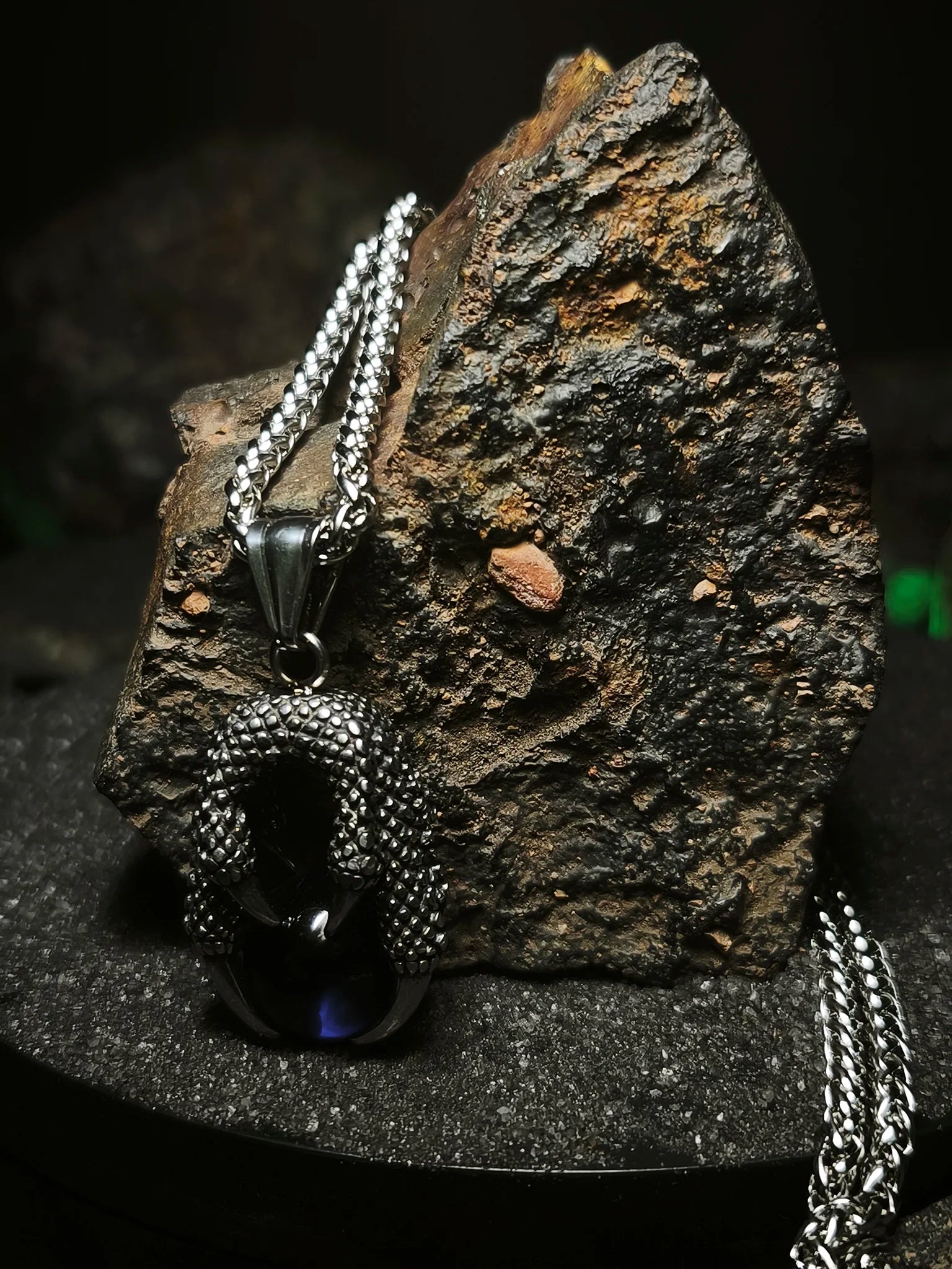 Orbe del Dragón Azul: Detalle de cadena de acero inoxidable macizo con colgante de garra y orbe místico sobre roca volcánica en Chile.